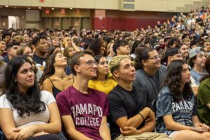 A large group of diverse students sit closely together in a gymnasium, attentively watching an event. Some wear shirts with Ramapo College printed on them. The atmosphere appears lively and engaged.