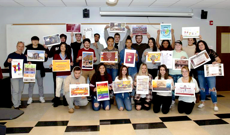 A group of students and a teacher stand in a classroom, holding up colorful posters and projects, smiling at the camera. The classroom has a whiteboard in the background and desks in the foreground.