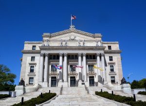 A grand neoclassical courthouse with tall columns, statues, and three flags on the steps, including the American flag. The building stands under a clear blue sky.