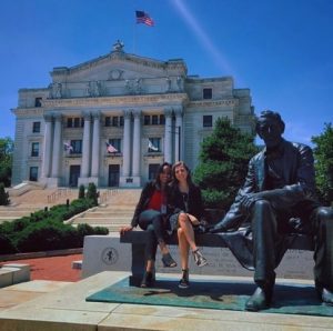 Two women sit on a bench next to a bronze statue in front of a large white courthouse with columns and an American flag flying on top, under a clear blue sky.