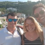 Three people smiling for a selfie outdoors near a harbor, with hills, boats, and buildings in the background. Two men wear sunglasses and white shirts; the woman in the middle has long blonde hair and is wearing a striped dress.