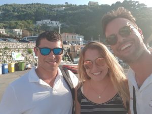 Three people smiling for a selfie outdoors near a harbor, with hills, boats, and buildings in the background. Two men wear sunglasses and white shirts; the woman in the middle has long blonde hair and is wearing a striped dress.
