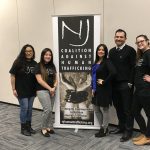 Five people stand smiling next to a banner that reads NJ Coalition Against Human Trafficking, with interlocked hands pictured, in a conference room setting. The groups website is listed at the bottom of the banner.