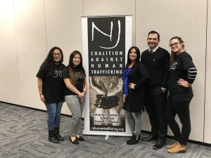Five people stand smiling next to a banner that reads NJ Coalition Against Human Trafficking, with interlocked hands pictured, in a conference room setting. The groups website is listed at the bottom of the banner.