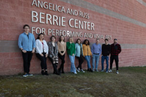 Ten people stand in a row, smiling, in front of the brick wall with the large sign Angelica and Russ Berrie Center for Performing and Visual Arts, home to Student Galleries. The group appears relaxed and casually dressed.