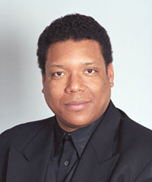 A man with short curly hair wearing a black suit and a black shirt, facing the camera and smiling slightly against a light gray background.