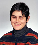 A person with short dark hair, wearing a gray turtleneck and a striped sweater with red, orange, and blue colors, smiles at the camera against a plain light background.