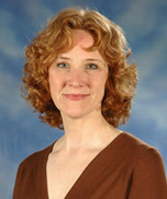 A woman with short, curly red hair and light skin smiles gently while wearing a brown top, standing in front of a blue and white blurred background.