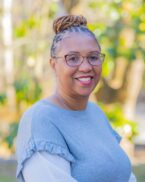 A woman with glasses and braided hair in a bun smiles while wearing a light gray top, standing outdoors with green and yellow blurred foliage in the background.