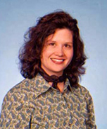 A woman with wavy brown hair, wearing a patterned shirt and a dark scarf around her neck, smiles in front of a light blue background.