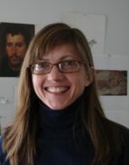 A woman with straight light brown hair and glasses smiles at the camera, wearing a dark turtleneck. Behind her, there are several artworks and a portrait on a white wall.