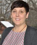 A person with short dark hair, wearing a gray blazer over a patterned shirt, stands outdoors with trees and a wooden structure in the background.