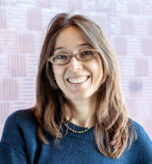 A woman with long brown hair and glasses smiles at the camera. She is wearing a dark blue sweater and a beaded necklace. The background is a soft purple mosaic tile pattern.