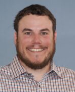 A man with short brown hair and a beard, smiling at the camera. He is wearing a plaid shirt with a blue background.