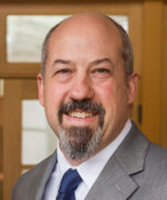 A middle-aged man with a bald head and gray goatee, wearing a gray suit, white shirt, and navy tie, is smiling in front of a wooden door with glass panels.
