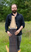 A person with a beard stands outdoors on grass, holding a large, multi-stringed wooden instrument vertically in front of their body. Surrounded by trees and greenery, they appear immersed in the evolution of music technology.