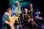 Four people in colorful, dramatic costumes gather around a serious, dark-clad figure. Theatrical lighting and props, including a tiered tray of pastries, set the scene—perfect for immersive Theater Productions.