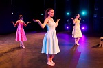 Three women in colorful dresses perform a dance routine on stage, each with arms raised and palms up. The stage, reminiscent of lively theater productions, is lit with vibrant blue and purple lights in the background.