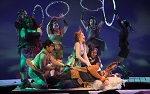 A group of actors in colorful costumes perform energetically on stage during one of the theater productions, some holding hoops above their heads, while two central figures interact dramatically under vibrant theatrical lighting.
