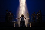A person in a long dress stands center stage with arms outstretched under a dramatic beam of light, surrounded by musicians against a dark blue background, capturing the magic and intensity of Theater Productions.
