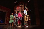 Five actors stand on stage in costume, lined up behind a microphone, performing in front of a large arched window set piece with dramatic lighting—a captivating scene from one of the theater productions.