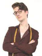 A person with short wavy hair and glasses stands with arms crossed, wearing a dark patterned shirt and a yellow measuring tape around their neck, against a plain white background.