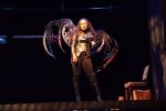 A performer in a metallic costume with large, wing-like extensions stands on a dark stage, illuminated by a spotlight—a striking scene often seen in innovative theater productions. A small wooden bench and desk sit quietly in the background.