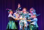 Five people in shiny, metallic costumes perform energetically on stage against a purple curtain, capturing the excitement of Theater Productions as one person in the center poses dramatically while others surround and reach toward them.