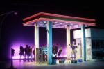 A group of people dancing and raising their arms under a brightly lit, retro-style gas station canopy at night evokes the energy of Theater Productions, with a purple-lit background creating a dramatic, theatrical scene.