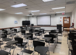 A 100 Classroom with showing desks throughout the room facing a projector screen 