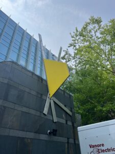 A large sculpture installed on the side of a glass and concrete building.The sculpture has intersecting metal rods and a large yellow metal centerpiece at the point where these rods intersect.