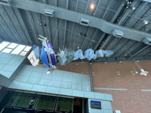 A sculpture suspended from a metal ceiling. The sculpture is made of twisted metal, mostly silver in color but with two large blue circular pieces on the front end.