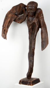 Wood carving of an upright figure with drooping wings. The clearest part of the figure is the face, with a kind expression and head wrap. The body is one continuous length of wood with no discernible limbs.