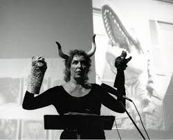 A person wearing horned headgear and animal-like gloves stands at a podium, with a large image of a dinosaur or monster displayed on a screen behind them. The scene appears theatrical and dramatic.
