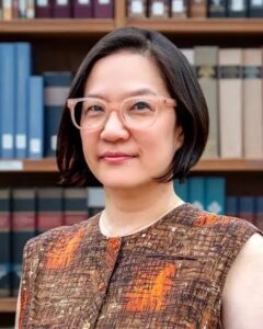 A woman with short dark hair and clear glasses wears a sleeveless brown patterned top, standing in front of bookshelves filled with books.