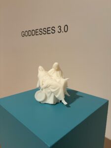 A small white sculpture of two seated, veiled female figures is displayed on a teal pedestal. Behind them, the wall text reads “GODDESSES 3.0” in bold black letters.