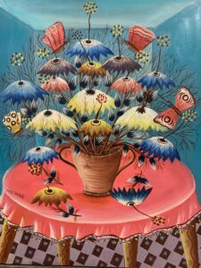 Upon a table with a bright pink tablecloth sits a large centerpiece with blue, yellow, pink, and purple umbrella-shaped flowers. Similarly colored butterlies rest on and around these flowers. The table is in a blue-walled room with a checkered floor.