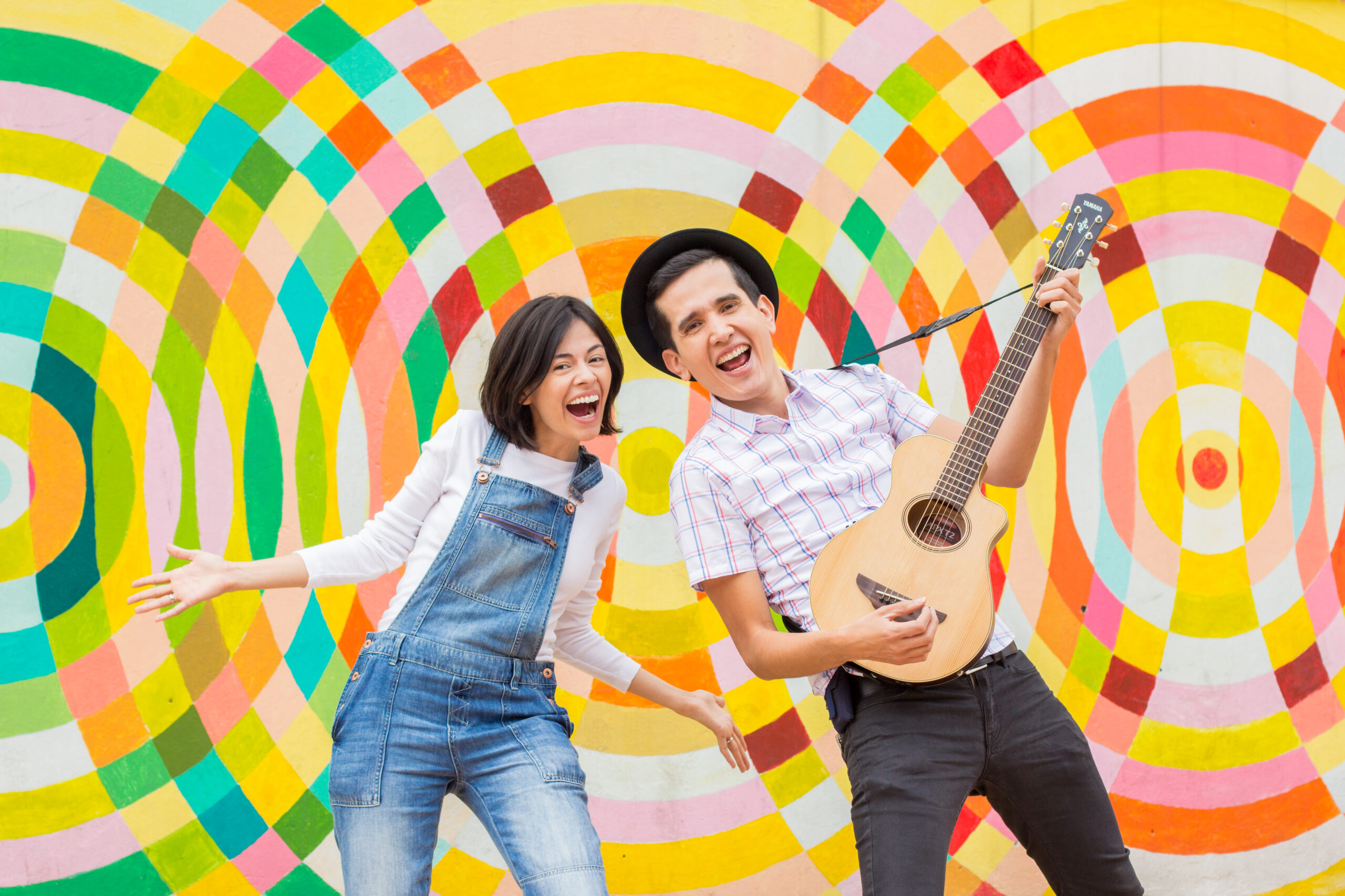 Andres and Christina in front of color background. Andres playing guitar