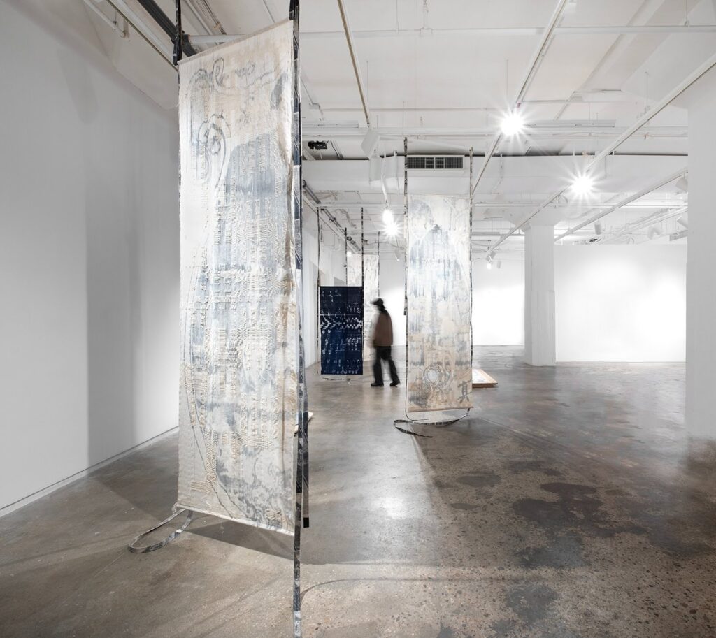 A gallery space with standing textiles, primarily white, and a blurry figure walking among them.