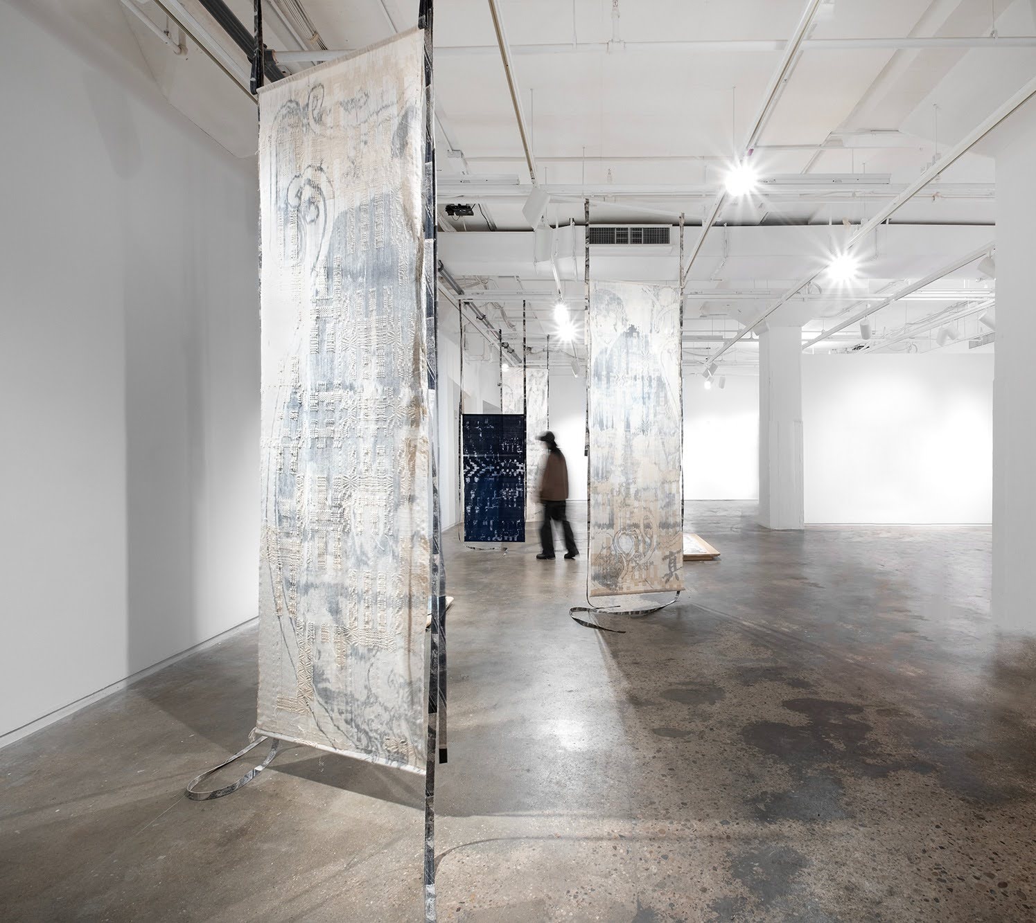 A gallery space with standing textiles, primarily white, and a blurry figure walking among them.