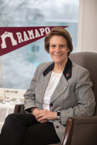 Ramapo College's President, Cindy Jebb, wearing plaid blazer sits in a chair, smiling, with a maroon Ramapo pennant displayed behind her and a window in the background.
