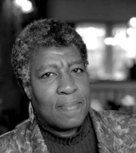 Black and white portrait of a middle-aged person with short curly hair, wearing a patterned jacket, earrings, and turtleneck sweater, looking thoughtfully at the camera with a softly blurred background.