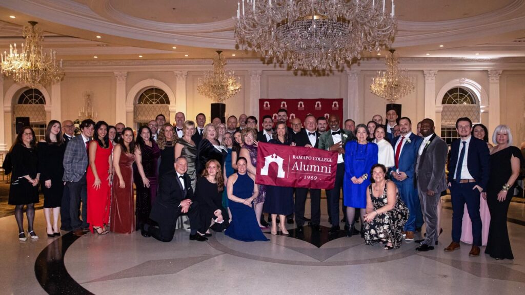 A large group of people dressed formally stand together in a grand ballroom, smiling and holding a red banner that reads “Ramapo College Alumni 1969.” Chandeliers and elegant decor illuminate the room.