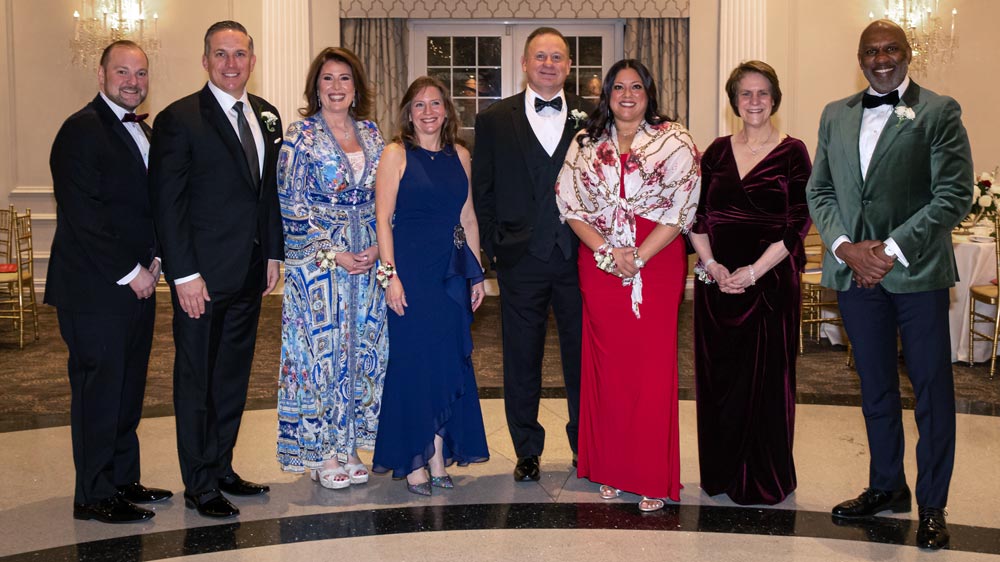 A group of eight adults dressed in formal evening attire stand in a row inside an elegant, well-lit banquet hall with chandeliers and gold chairs in the background.