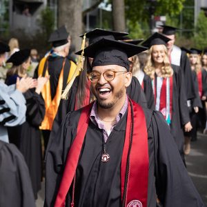 Student wearing glasses in regalia, surrounded by other students wearing regalia