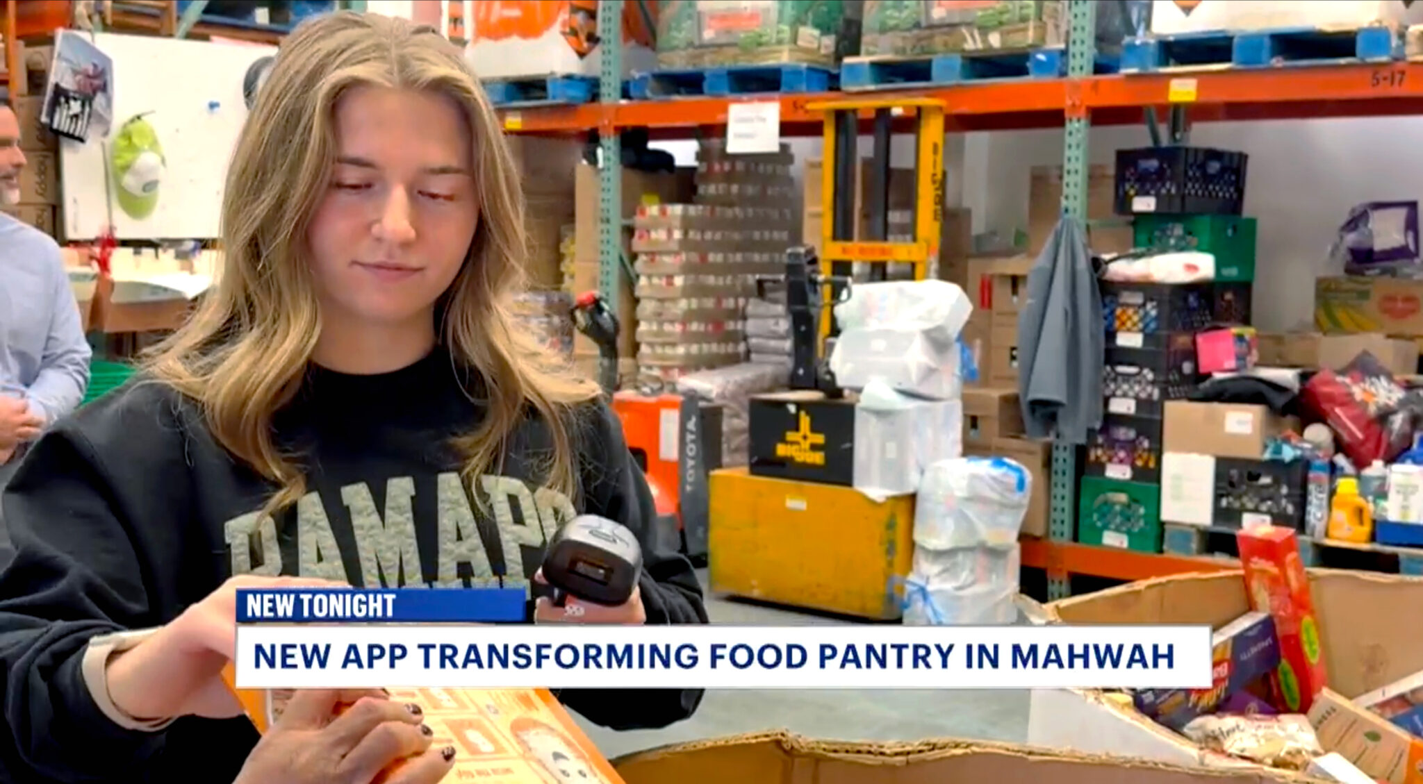 Student with long blond hair, wearing a dark Ramapo sweatshirt and holding a dark scan gun about to scan barcode of food box at the Center for Food Action (CFA) in Mahwah.