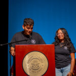 Two students stand at a podium with the Ramapo College of New Jersey seal, speaking at an event. They wear matching black shirts, and several people are seated in the background on stage.
