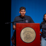 A young man and woman stand at a podium with the Ramapo College of New Jersey seal, speaking on stage. Both wear matching dark t-shirts, and the background is blue with other people seated behind them.