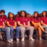 A diverse group of teens in matching maroon shirts sit on folding chairs on stage, facing forward. The background is a blue gradient, and the students appear attentive and engaged.