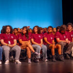 A large group of students in matching maroon t-shirts sit onstage in rows of chairs, facing forward. The background is a solid blue. The students appear attentive, with some looking at the camera.